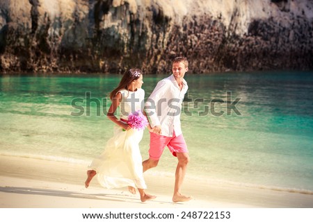 handsome groom run with his beautiful brunette bride in long white wedding dress on island bay with green mountain background in Thailand