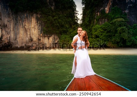handsome groom embrace blonde bride in wedding dress sitting on the nose of longtail boat near the rocky island