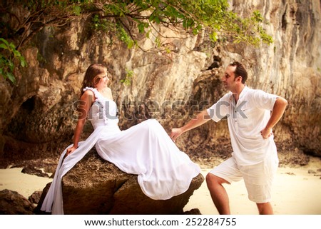 handsome groom looks at beautiful bride sitting on the rock at the sand beach