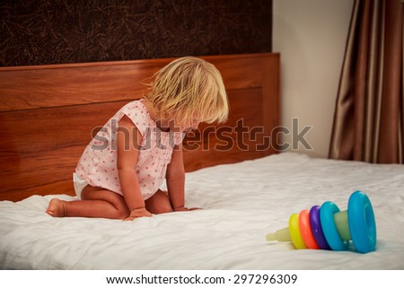 portrait of sitting on knees little blonde girl in pink dress near toy pyramid against brown wall