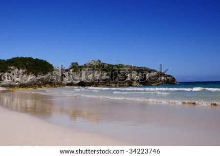Bermuda\'s Horseshoe Bay beach during a hot summer day.  Photo includes the sky, large rocks along the coastline, pink sand and the tide rolling in.