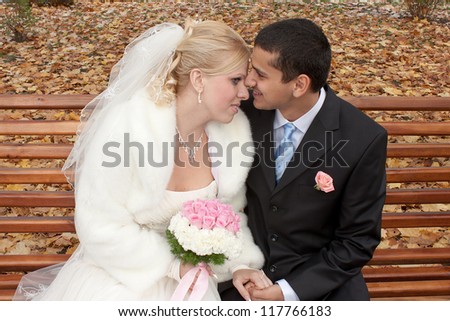 Happy bride and groom sitting on the bench, holding hands and smiling. Groom kissing bride.