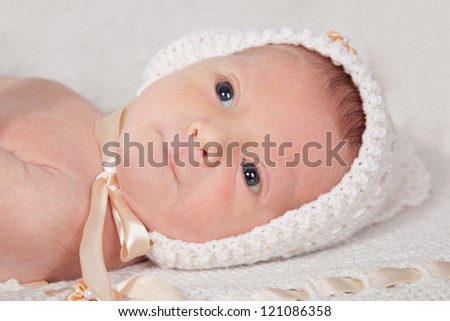 10 day old newborn baby in white fashion knitting hat laying on white knitting blanket looking curious and bite his lip. Selective focus on baby face
