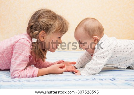Older sister holding hands of her little sibling. Children lying on their tummies on the bed. Both laughing and having fun together. Side view.