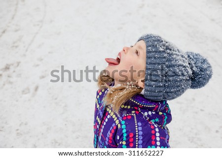 Little girl dressed in winter clothes catching snowflakes with her tongue while walking through falling snow. Winter fun. Close up.