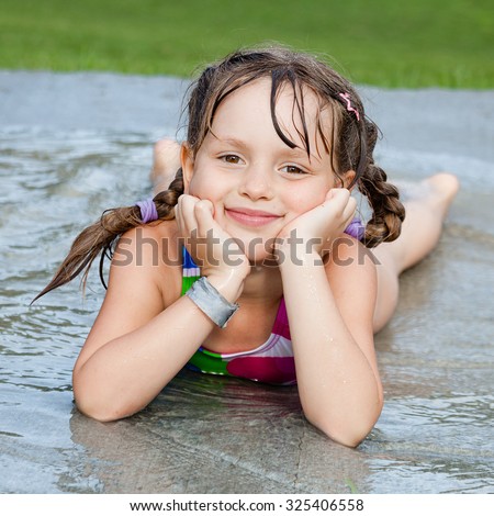 Little girl lying in the water stream on child splash pad playground. Summer fun while getting wet and refresh.