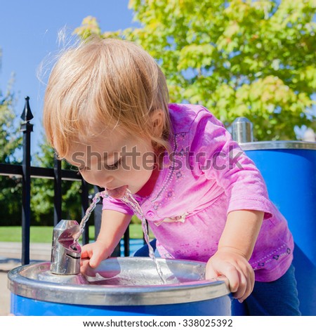 Cute baby drinking from water drinking fountain in summer. Toddler girl first attempts to do adult things. Bright sun, hard shadows. Selective focus on water stream and girls face.