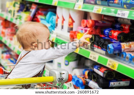 Cute baby in the supermarket trolley is reaching to shop stuff in household goods department
