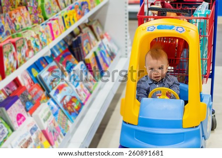 Happy baby boy in the little toy-car trolley in the kids shop in the bookstore department
