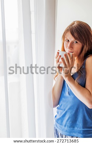 Beautiful redheaded girl with cup of tea or coffee near the window in the morning