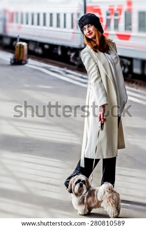 Elegant young woman in  hat with veil, with shih tzu dog and suitcase in the background. She is standing near train at the Vitebsk railway station in St.-Petersburg, Russia.