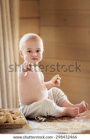Cute baby on the table smudgy with flour and homemade cookies in both hands. He is offering you to taste one. Image with selective focus