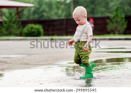 Cute little boy in cap and green rubber boots is having fun splashing through the puddles. Image with selective focus