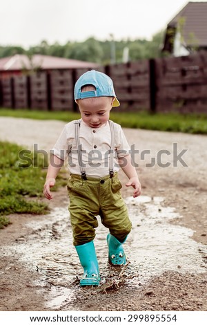 Cute little boy in cap and green rubber boots is having fun splashing through the puddles on the rural road. Image with selective focus