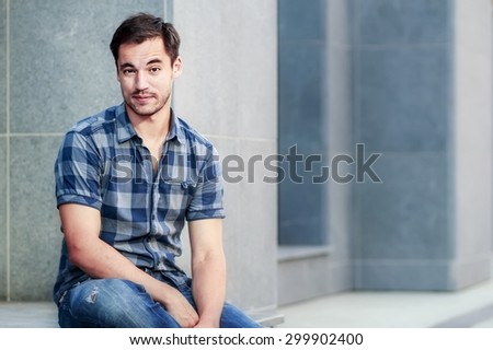 Portrait of attractive dark-haired young man with funny face expression sitting against an urban background. Image with selective focus