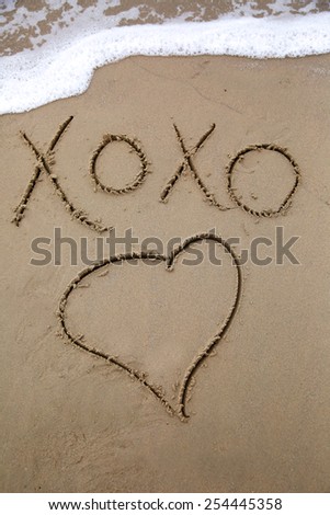 Message written in the sand kiss hug kiss hug and a drawing of a heart; message written in the sand about love with an ocean wave in the background with foamy bubbles