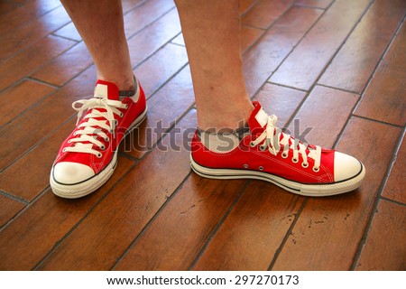 Red tennis shoes adorn a mans feet getting ready for a bug walk; red shoes and white shoelaces on a wood floor