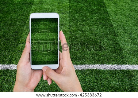 Hand holding mobile smart phone with football stadium, image of a football field as background.