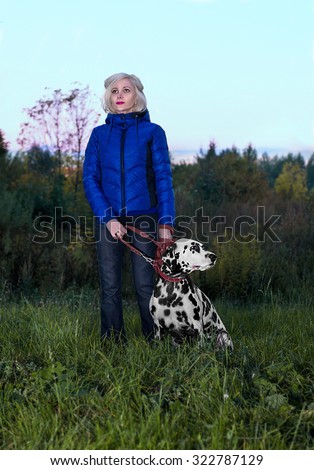 Beautiful blonde girl in a blue jacket and black jeans walking in nature with a splendid dog breed Dalmatian evening in the fall and they are looking into the distance