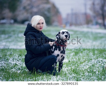 A young girl in a black warm jacket and black jeans hugging a Dalmatian dog, and sits with him on the first snow mixed with the green grass in the city and smile