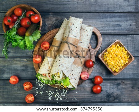 Sandwiches twisted roll Tortilla burritos three pieces on a wooden cutting board on a gray background, lettuce, cucumbers malosollnye, cherry tomatoes, corn, onion rings, meat, top view