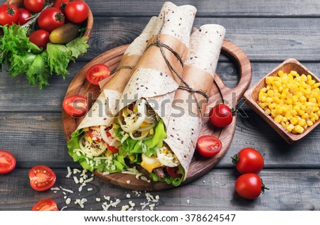 Sandwiches twisted roll Tortilla three pieces on a wooden cutting board on a gray background, lettuce, cucumbers malosollnye, cherry tomatoes, corn, onion rings, meat