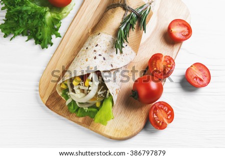Burrito wrapped in tortilla bread with cereals, corn, onion rings, lettuce, cheese, rosemary, pickled cucumbers, ketchup on a chopping board on a light white wooden background, top view