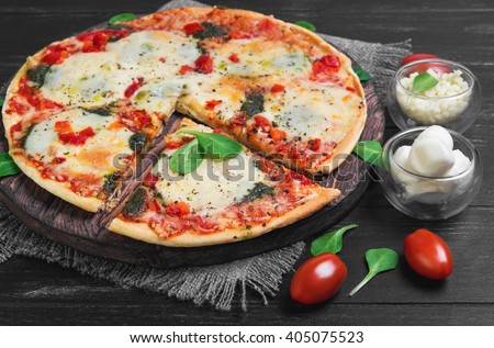 Pizza with mozzarella into balls and shredded cheese, cherry tomatoes,  cut off slice, lettuce on a cutting board round served on a dark black background wooden surface