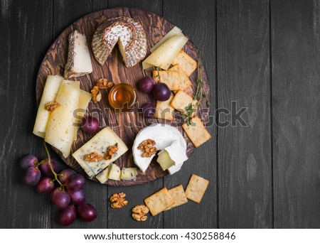 Assorted cheeses on round wooden board plate Camembert cheese, cheese grated bark of oak, hard cheese slices, walnuts, grapes, crackers, bread, thyme, dark black wood background, top view
