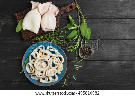 Boiled calamari squid rings on blue ceramic plate, cooked squid on cutting rings on board. Herbs for calamari squid rings thyme, sage, rosemary, black wooden background. Top view