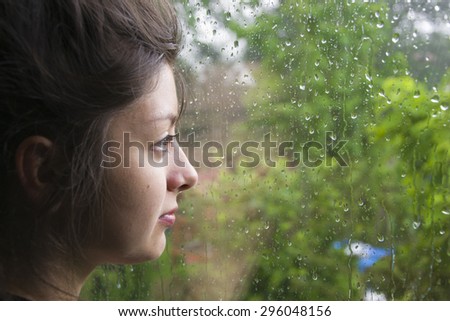beautiful young woman of Asian appearance looks out the window as the rain falls. The window in rain drops with green trees as background. Sadness and search. The girl has big black eyes.