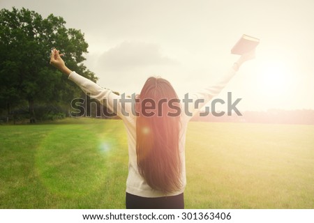 Beautiful woman with Bible is in sunny nature. Young lady meditate in the green forest with sun lights background.