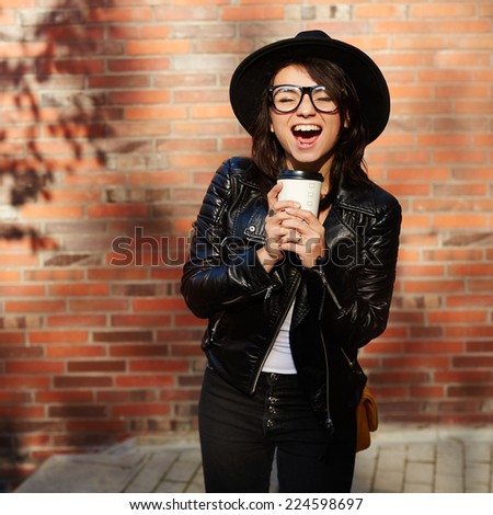 Portrait of a beautiful girl in a hat with a glass of coffee smiling on the background of the street. Hipster style.