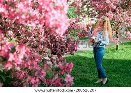 Girl photographs on a mobile phone flowering trees. Beautiful young girl with curly hair smiling on the background of an apple orchard. Fresh spring day.