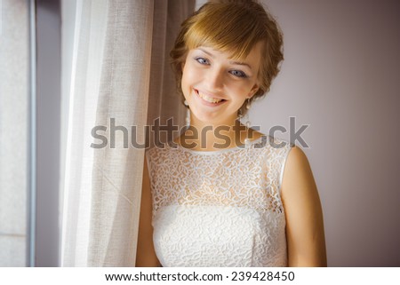 A young and beautiful bride standing by the window, smiling, waiting. A caucasian girl wearing a wedding dress.
