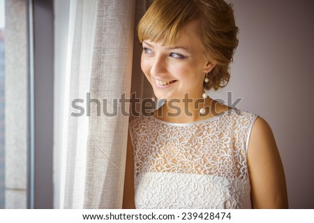 A young and beautiful bride standing by the window, smiling, waiting. A caucasian girl wearing a wedding dress. Smiling as she is look outside.