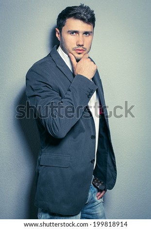 Fashion portrait of young handsome man wearing suit, jeans and white shirt posing over gray wall. Studio shot