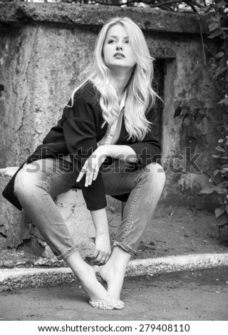 Sexy blond woman in trendy jeans and black jacket sitting barefoot. Street shooting. Vogue style.