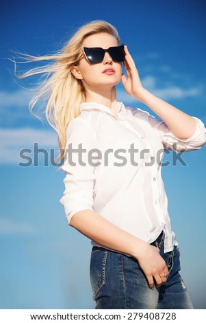 beautiful young woman in white shirt, jeans ad trendy sunglasses over blue sky.