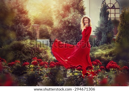 Romantic portrait of beautiful woman in fluttering red dress standing in the garden full of roses. Vintage photo.