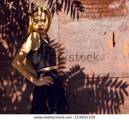 Light & Shadow fashion concept. Fashion portrait of beautiful blond-haired girl in trendy black crop top and skirt posing over rusty metal background. Perfect skin and make-up. Outdoor shot