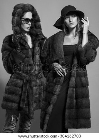 Fashion shot of two elegant beautiful girls (brunette and blonde) in studio on grey background wearing sunglasses, black hat and furs coat . Shopping inspiration. Monochrome (black and white)  photo