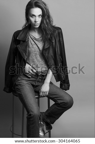 Portrait of a fashionable model with natural make up, perfect skin, dressed in men\'s jeans, grey shirt, black jacket and sneakers. Studio shot. High fashion look. Monochrome (black and white)  photo