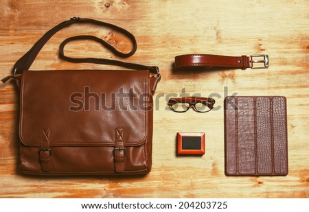 Male accessories style concent. Brown leather bag, box with french cuffs, trendy wooden rim glasses, leather belt and gadget in brown leather crocodile-textured case over wooden background. Copy space