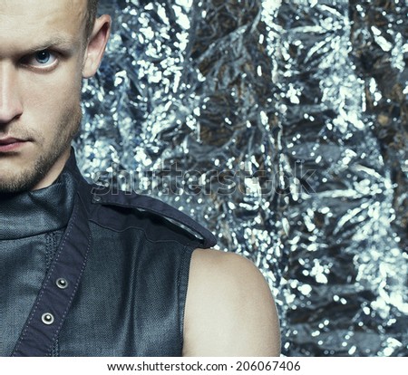 Young handsome blue-eyed man with strong intense look in sleeveless zip-front vest of dark jeans posing over silver foil background. Half-length portrait. Close-up. Studio shot