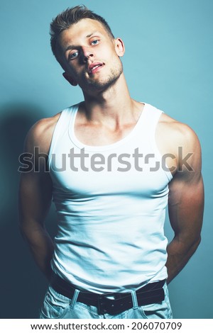 Handsome muscular blonde young man in white undershirt, light jeans and dark brown leather belt with seductive, sexy glass over blue background. Close up. Studio shot.