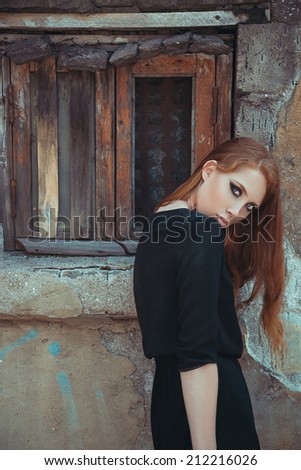 Old desert city fashion concept. Portrait of young beautiful red-haired girl in trendy black dress posing alone over old dream-window wall. Perfect skin and strong smoky eyes make-up. Hipster style.