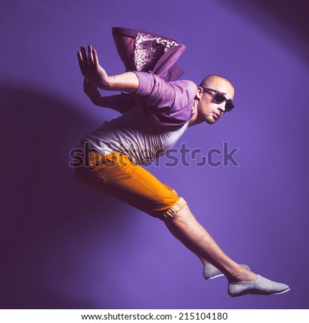 Young handsome male dancer in trendy glasses, purple jacket, white undervest and orange briefs jumps over gold background. Studio shot. Copy space.