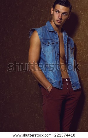 Male beauty concept. Portrait of handsome muscular male model in red pants and jeans west posing over gold background. Hand in pocket. Smooth head, healthy clean skin. Copy-space. Studio shot
