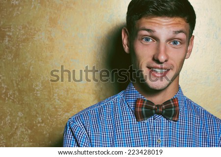 Emotive portrait of smiling handsome young man in blue plaid shirt and Scottish-plaid  bow-tie posing over golden background. White shiny smile and healthy skin. Hipster style. Studio shot. Copy-space